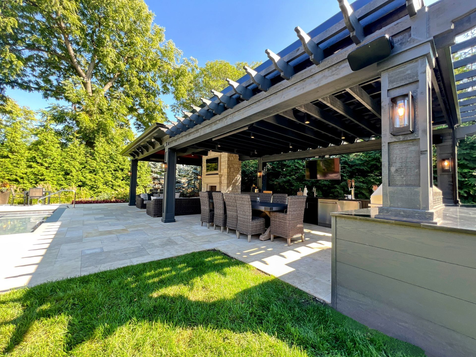 Patio with dark pergola, dining set, and built-in outdoor kitchen. Green lawn and trees surround the space.