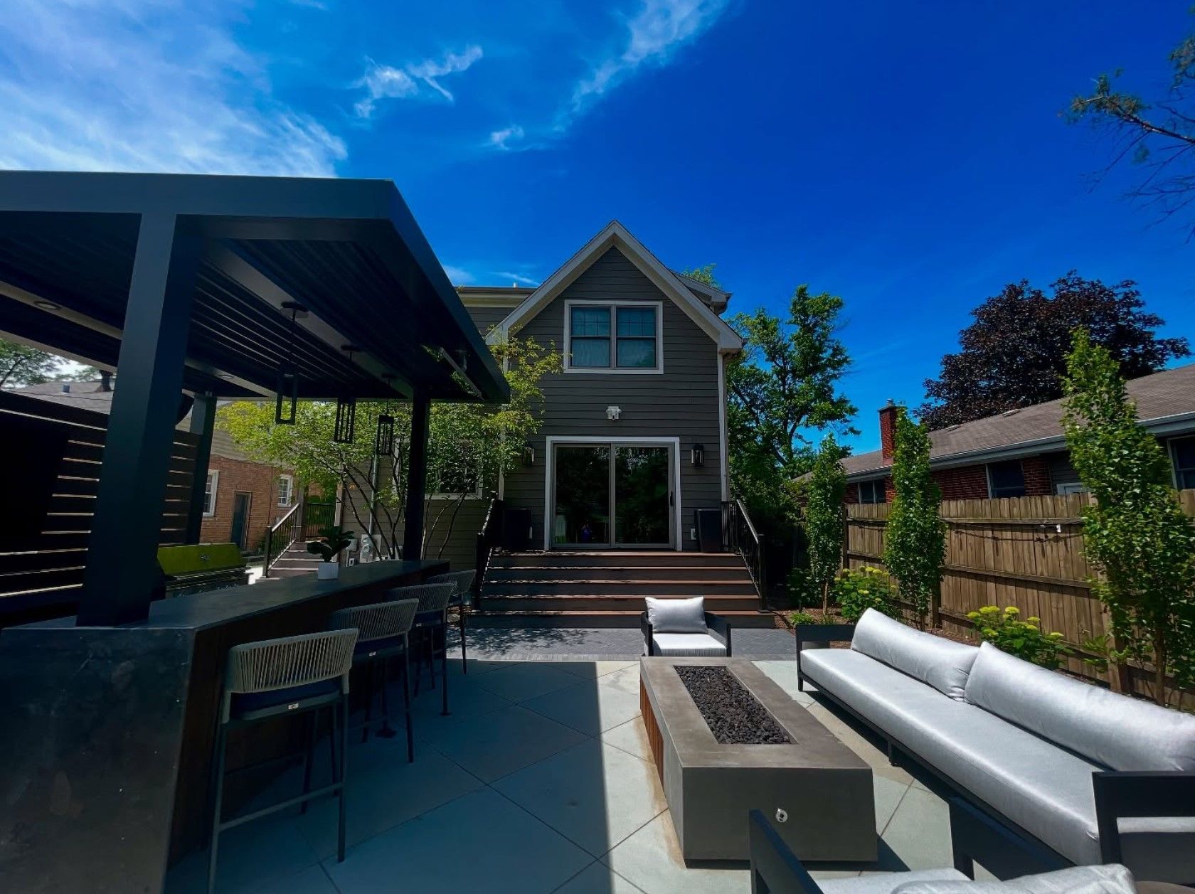 Backyard patio with bar, fire pit, and seating in front of a house, under a blue sky.