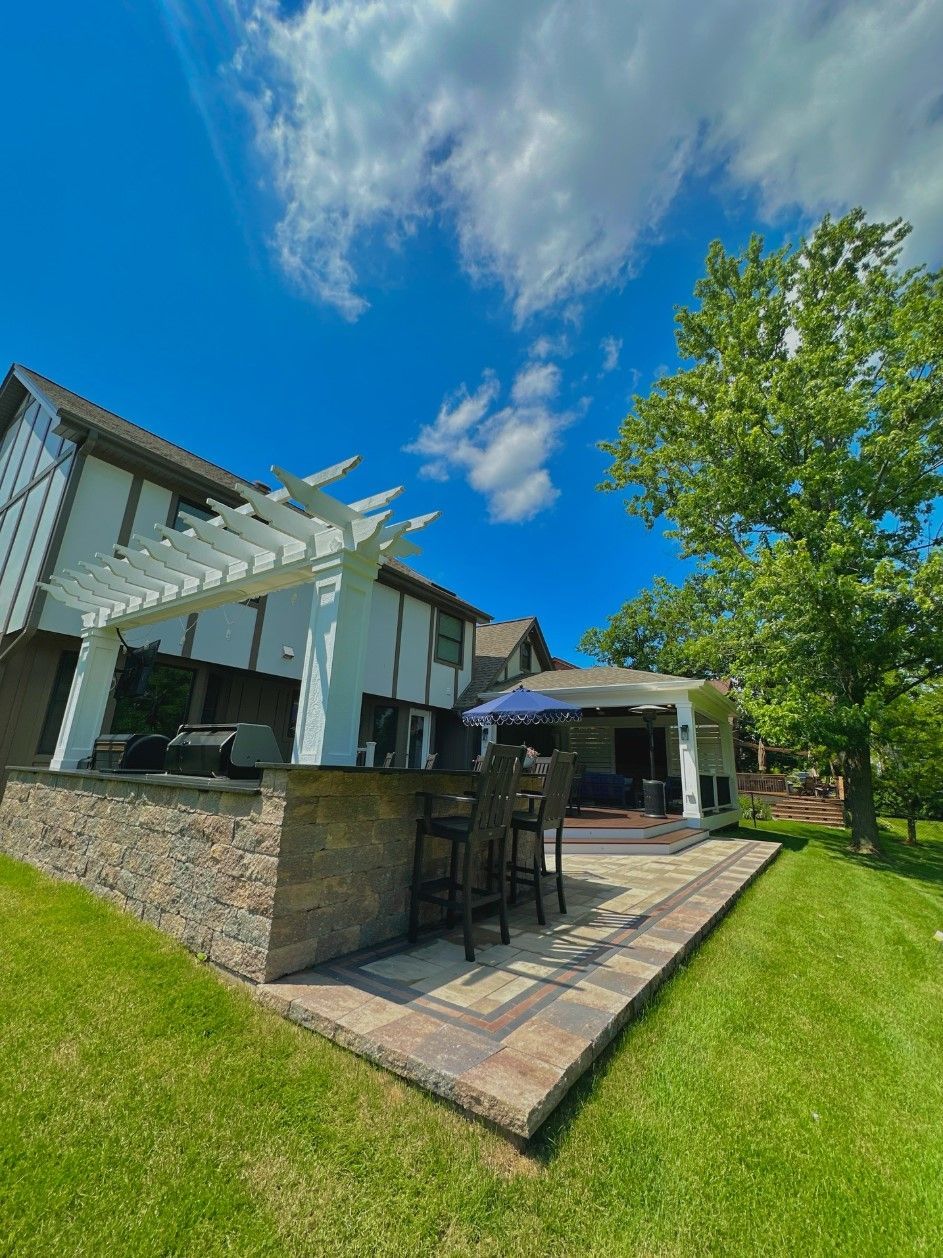 Patio with built-in bar, white pergola, house, and lush green lawn under a blue sky with clouds.