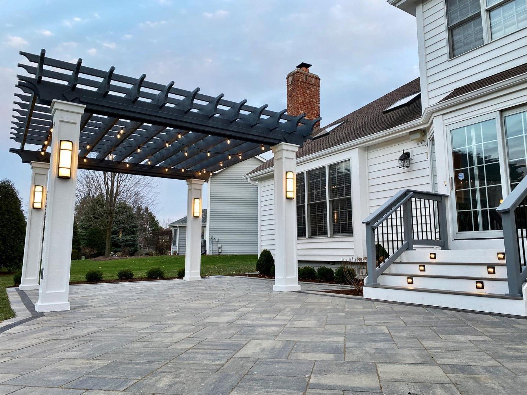 Pergola with string lights over patio, attached to white house with steps.