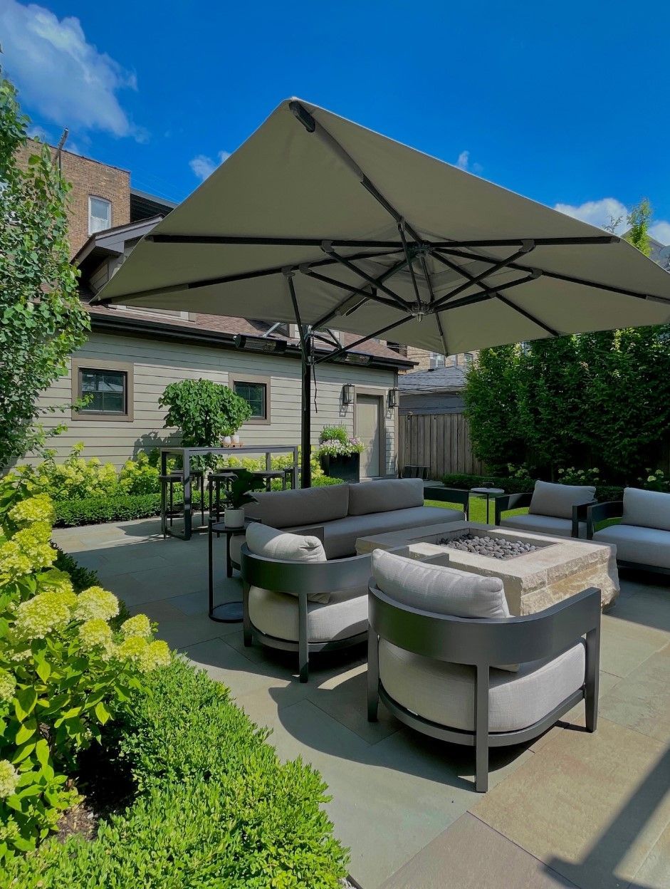 Patio with modern outdoor furniture, large umbrella, and lush greenery on a sunny day.