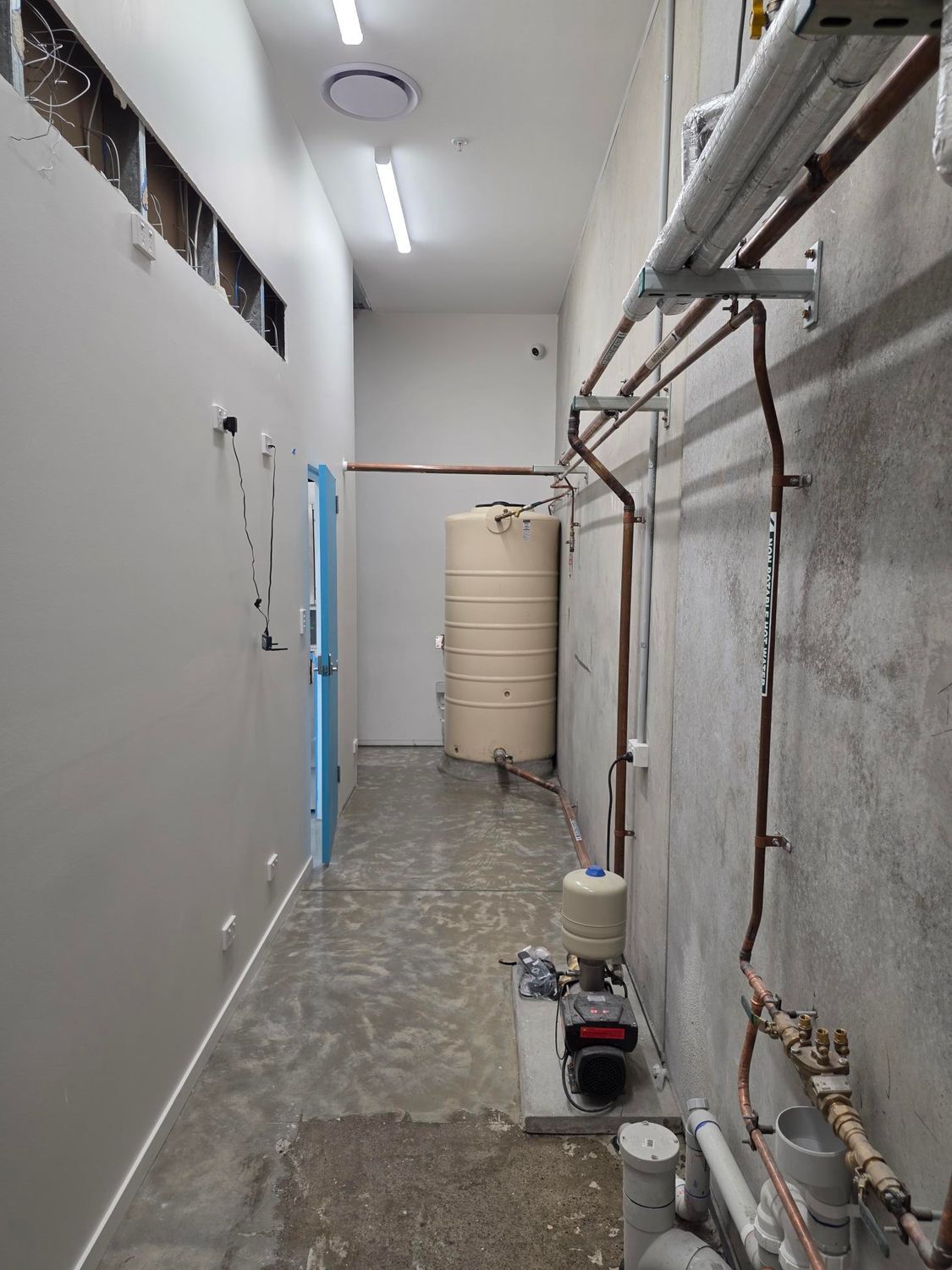 Narrow utility corridor with exposed pipes, white walls, and equipment on the floor.