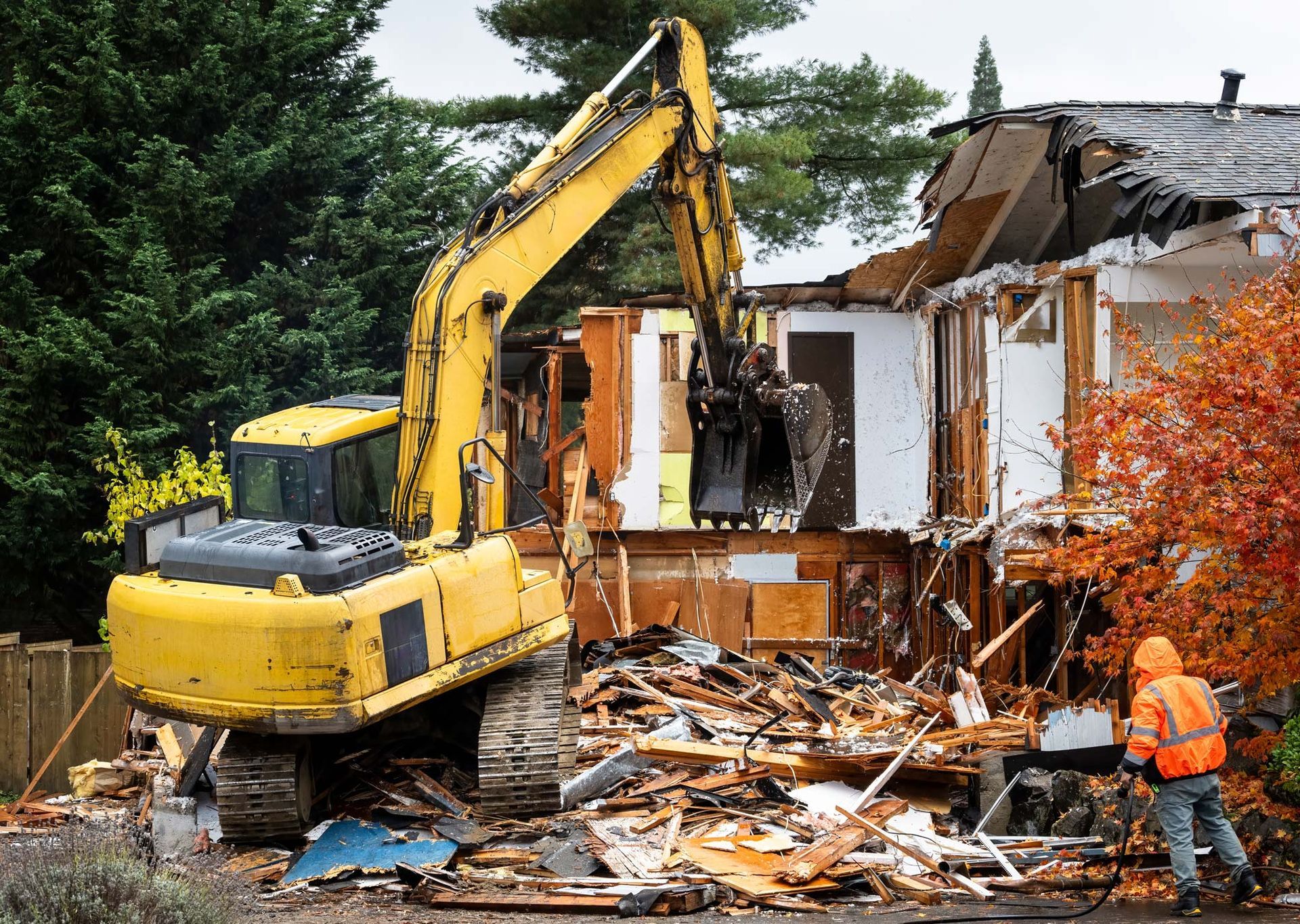 Yellow excavator demolishes a house; construction worker nearby.