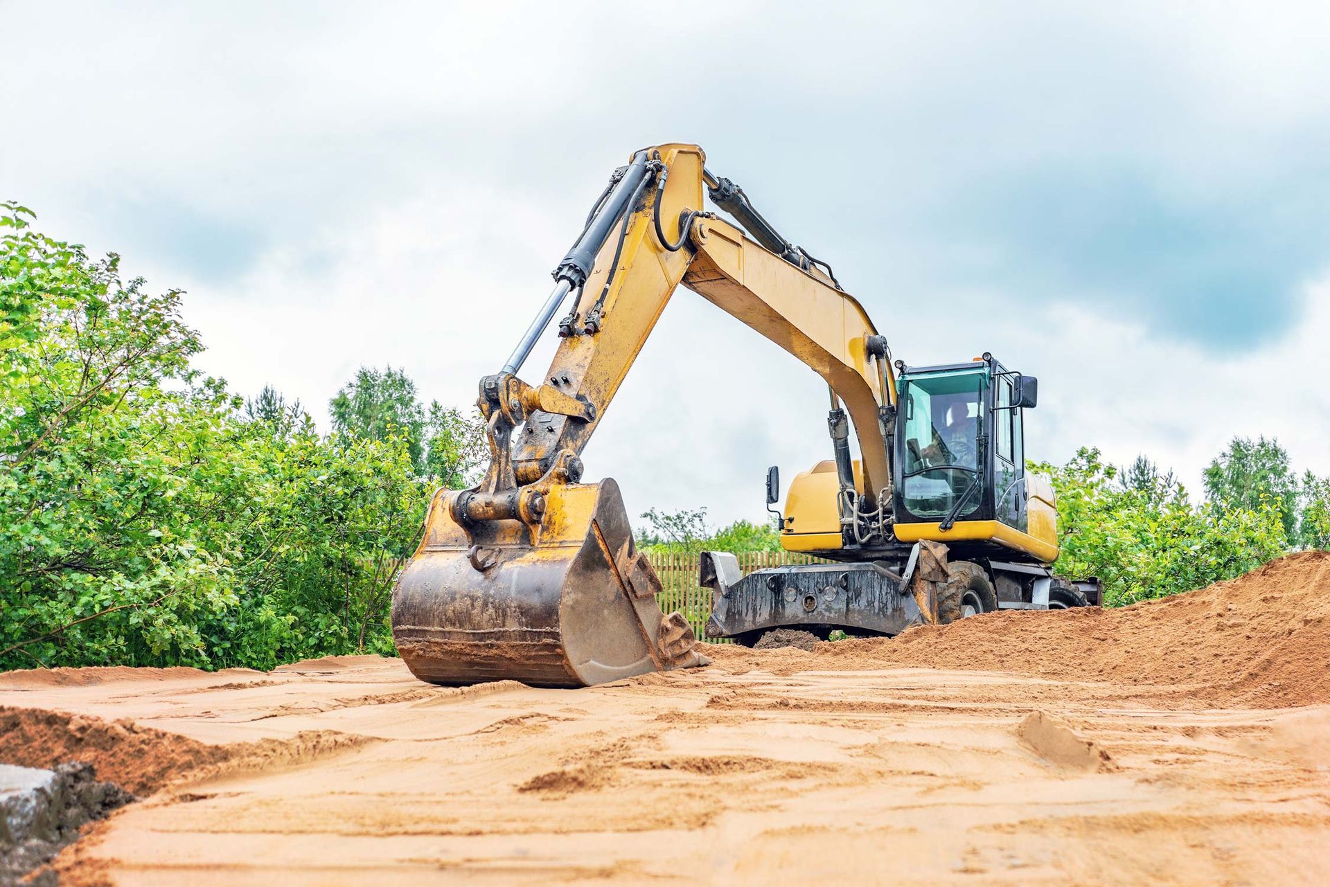 Yellow excavator digging in a dirt area, with trees in the background under a cloudy sky.