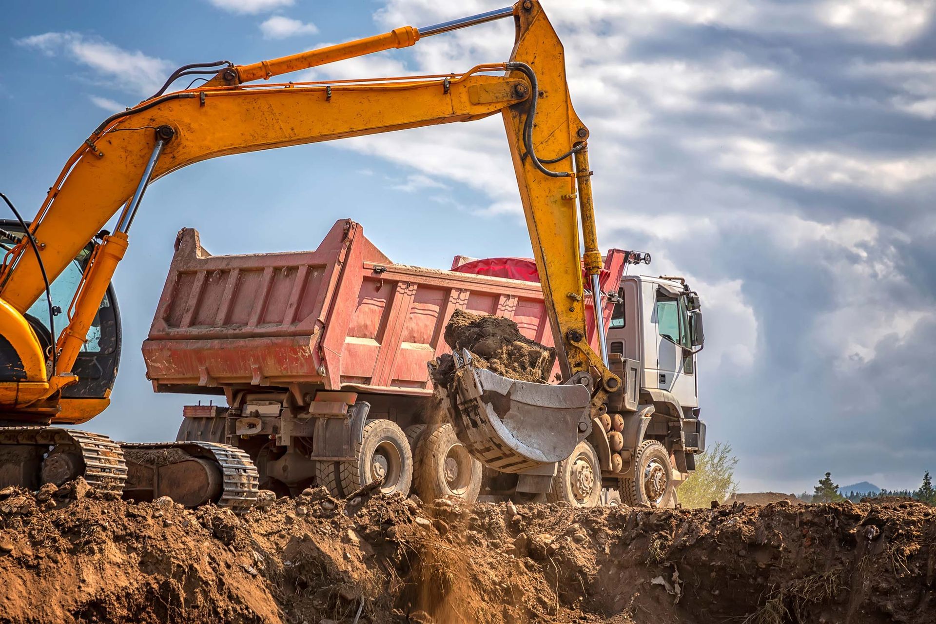 Yellow excavator loading a red dump truck with dirt on a construction site under a cloudy sky.