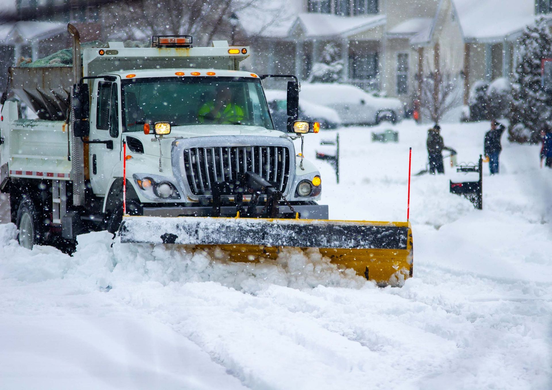 Snow plow clearing a snow-covered suburban street. Yellow blade pushes snow; white truck and houses in the background.
