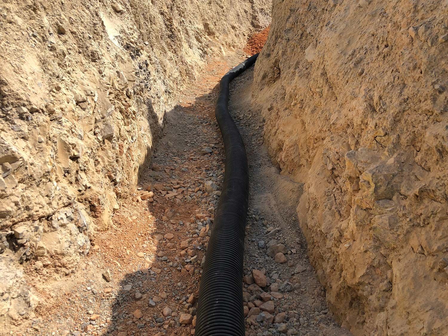 A black corrugated drainage pipe installed in a narrow trench between two dirt walls.