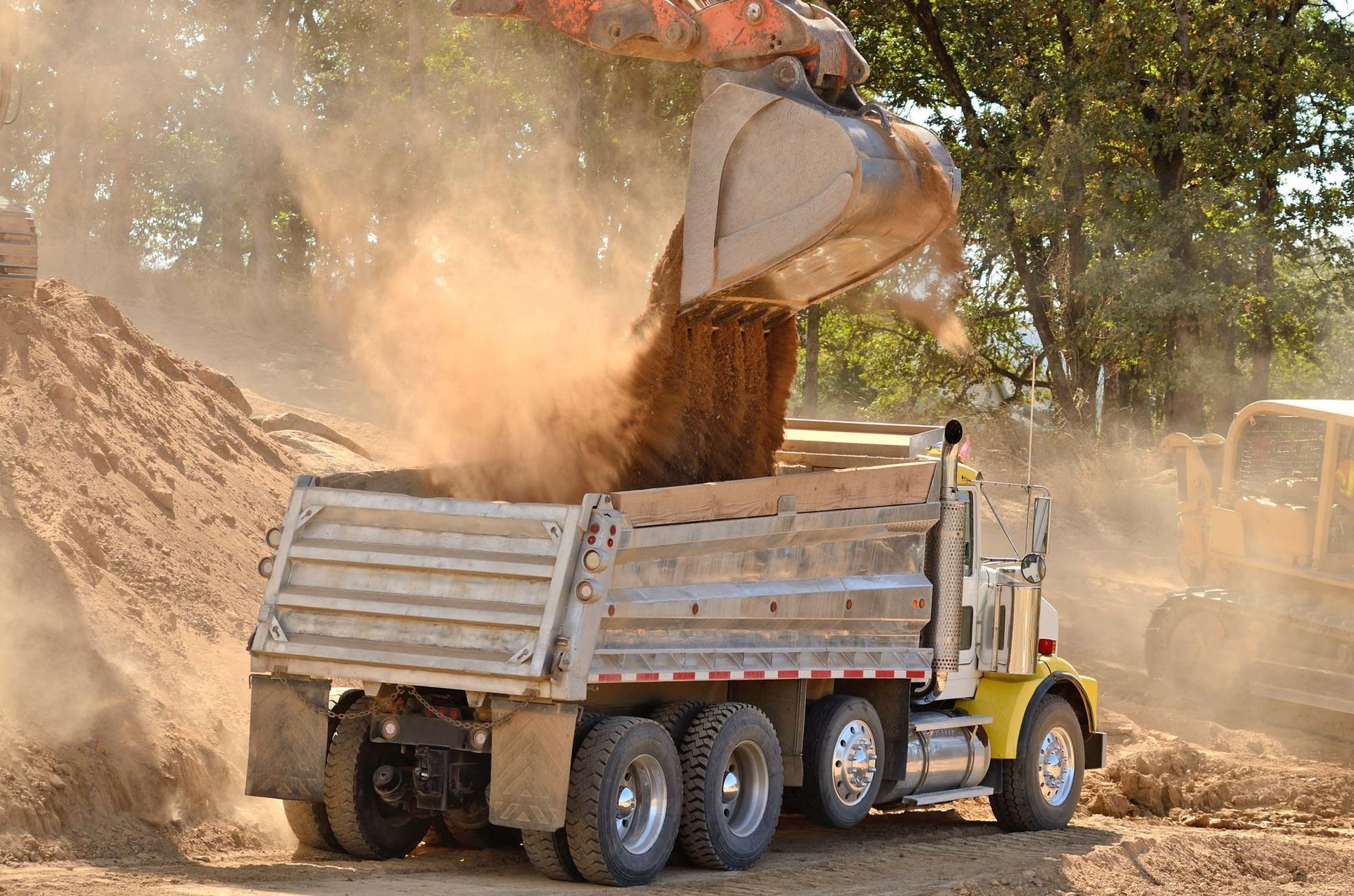 An excavator loading brown dirt into a yellow dump truck at a construction site.