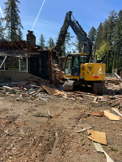 Yellow excavator demolishes a house; construction worker nearby.