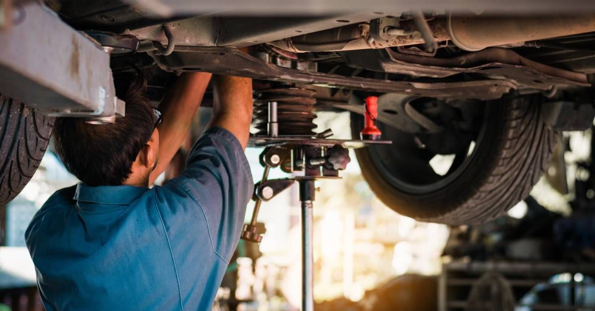 A mechanic wearing a blue shirt and glasses works on a suspension system underneath a lifted car.