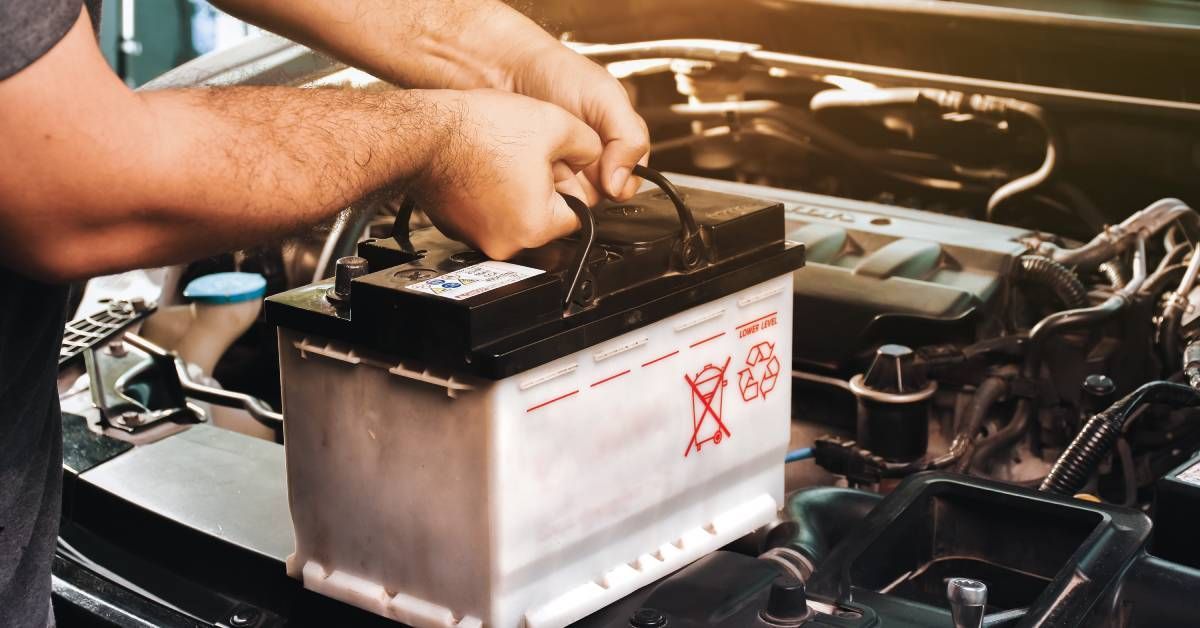 An auto mechanic holds a car battery on top of a car's engine.