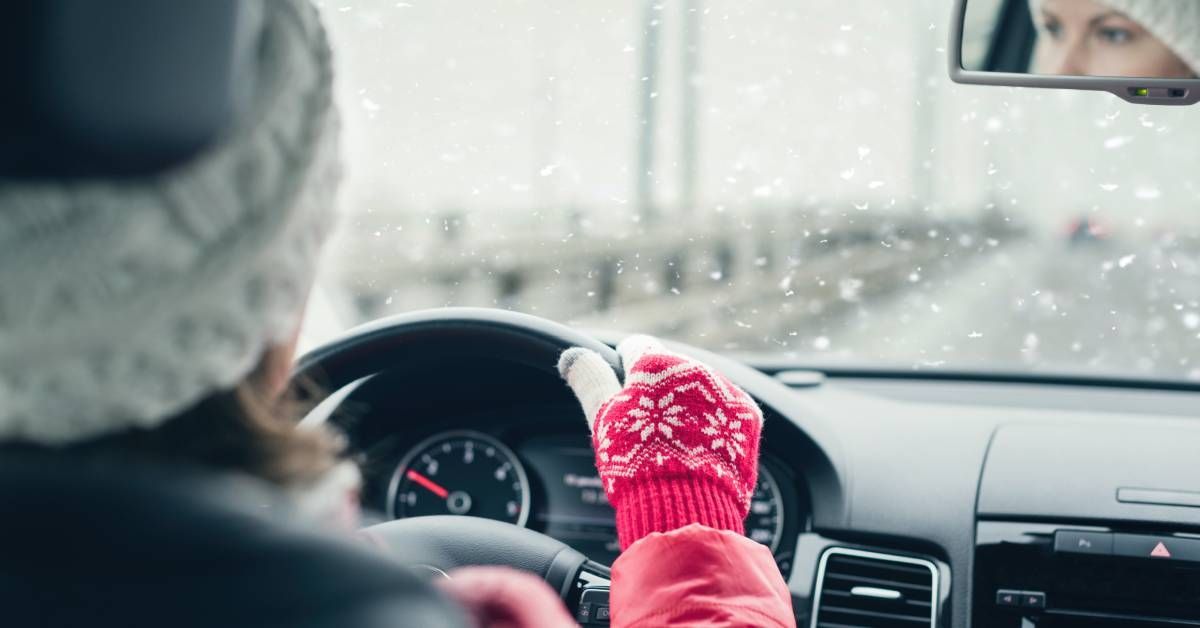 A woman drives her car while the snow falls. She has a winter hat and gloves on.