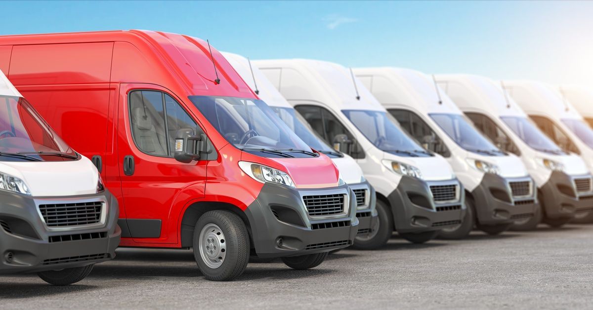 A red van is parked beside a row of white vans, all beneath a bright blue sky.