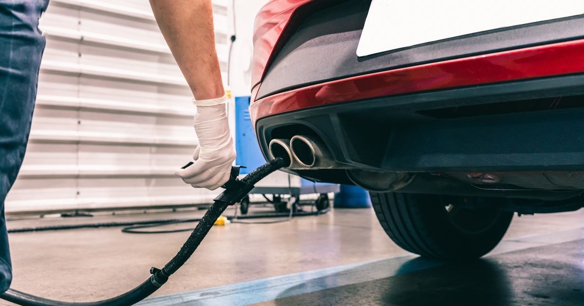 A technician wearing white gloves performs an emissions test check using a scanner on a red vehicle.