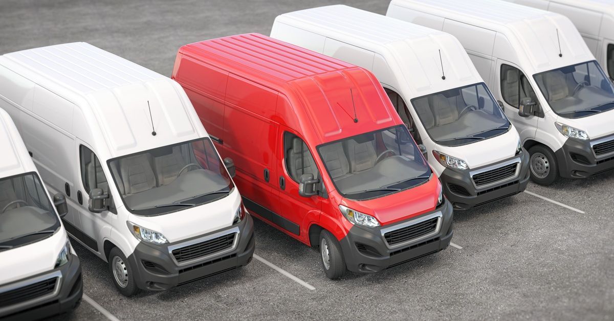 A red delivery van stands out is parked in a lot among a fleet of white vans.