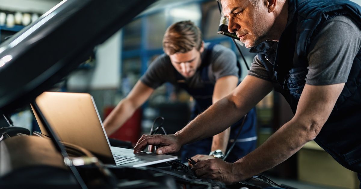 Two mechanics leans and works on getting a car diagnostic using a laptop.