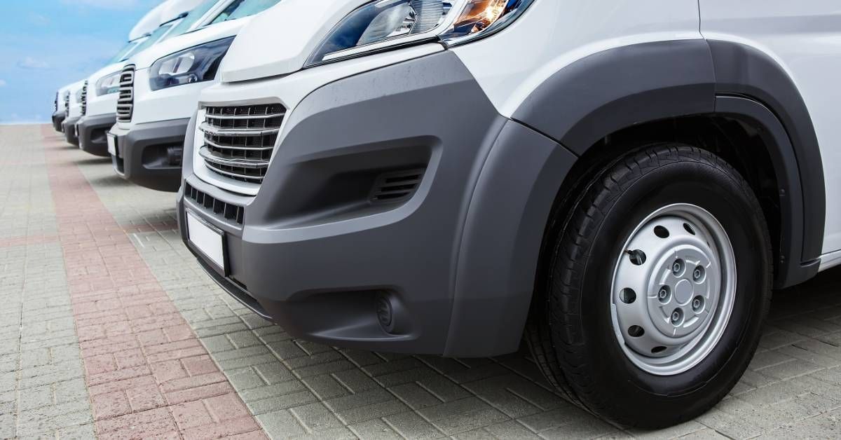 Minibuses and vans parked outdoors in a row in a parking lot, trees behind.