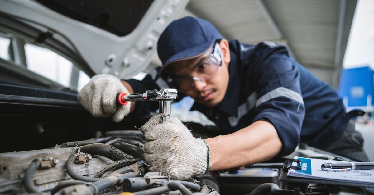 A mechanic in a blue uniform leans over a car's engine and completes work with a ratchet.