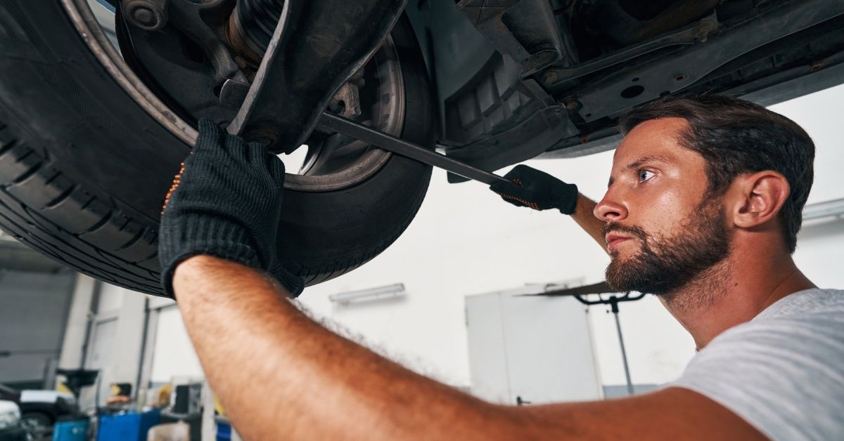A man wearing a white shirt uses a crow bar on a car's suspension system.