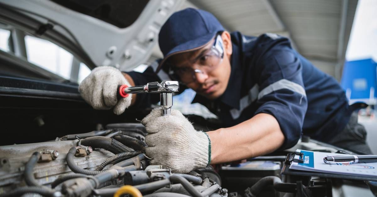 A man wearing safety glasses uses a ratchet on a car engine beneath an open hood in a vehicle garage