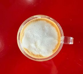 A cup of coffee on a red saucer next to a pile of coffee beans.