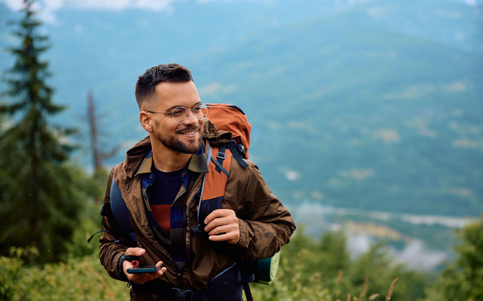 Man with backpack smiles while hiking in mountains, holding a phone.
