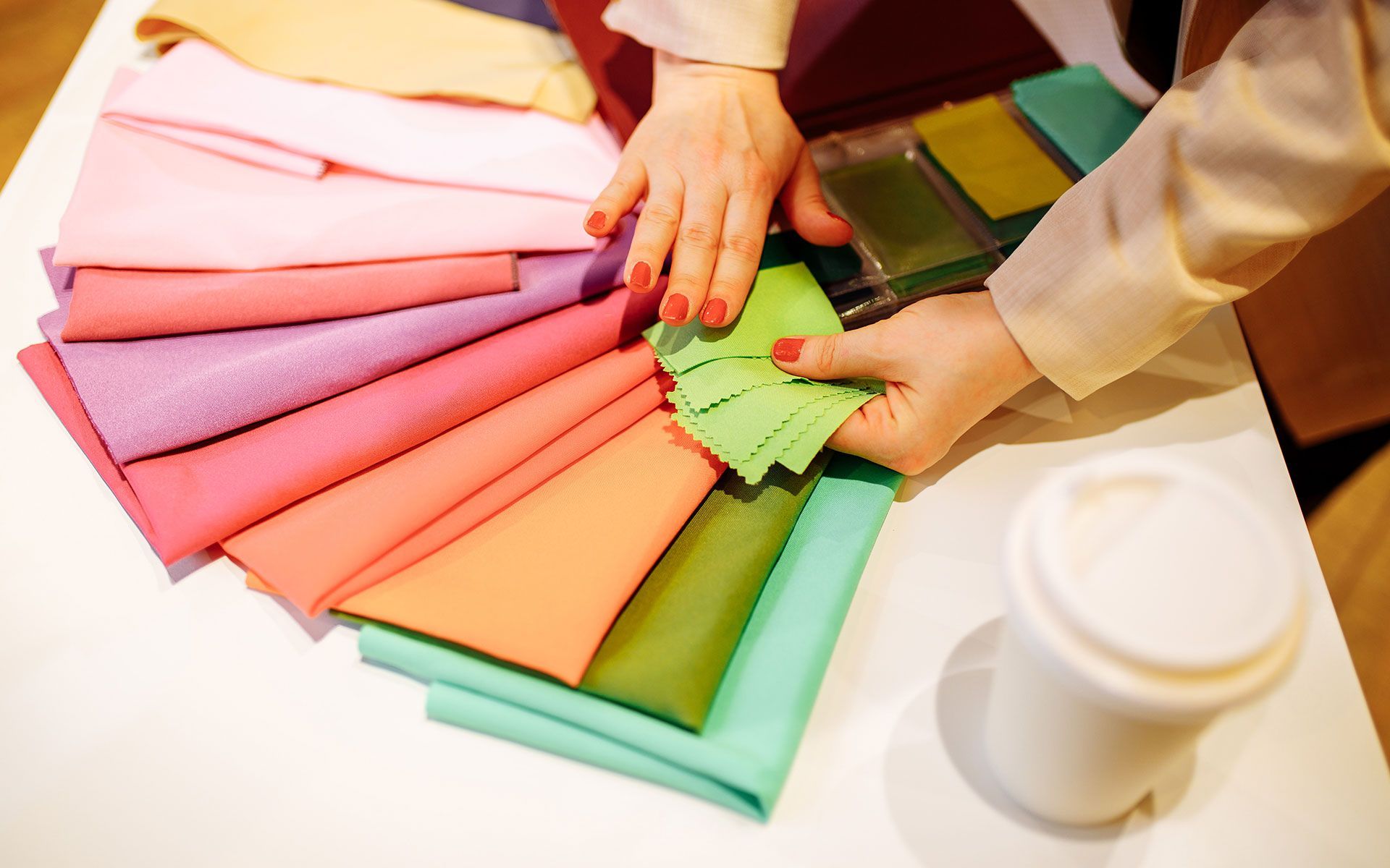 Hands arranging colorful fabric swatches on a white surface, with a coffee cup on the side.
