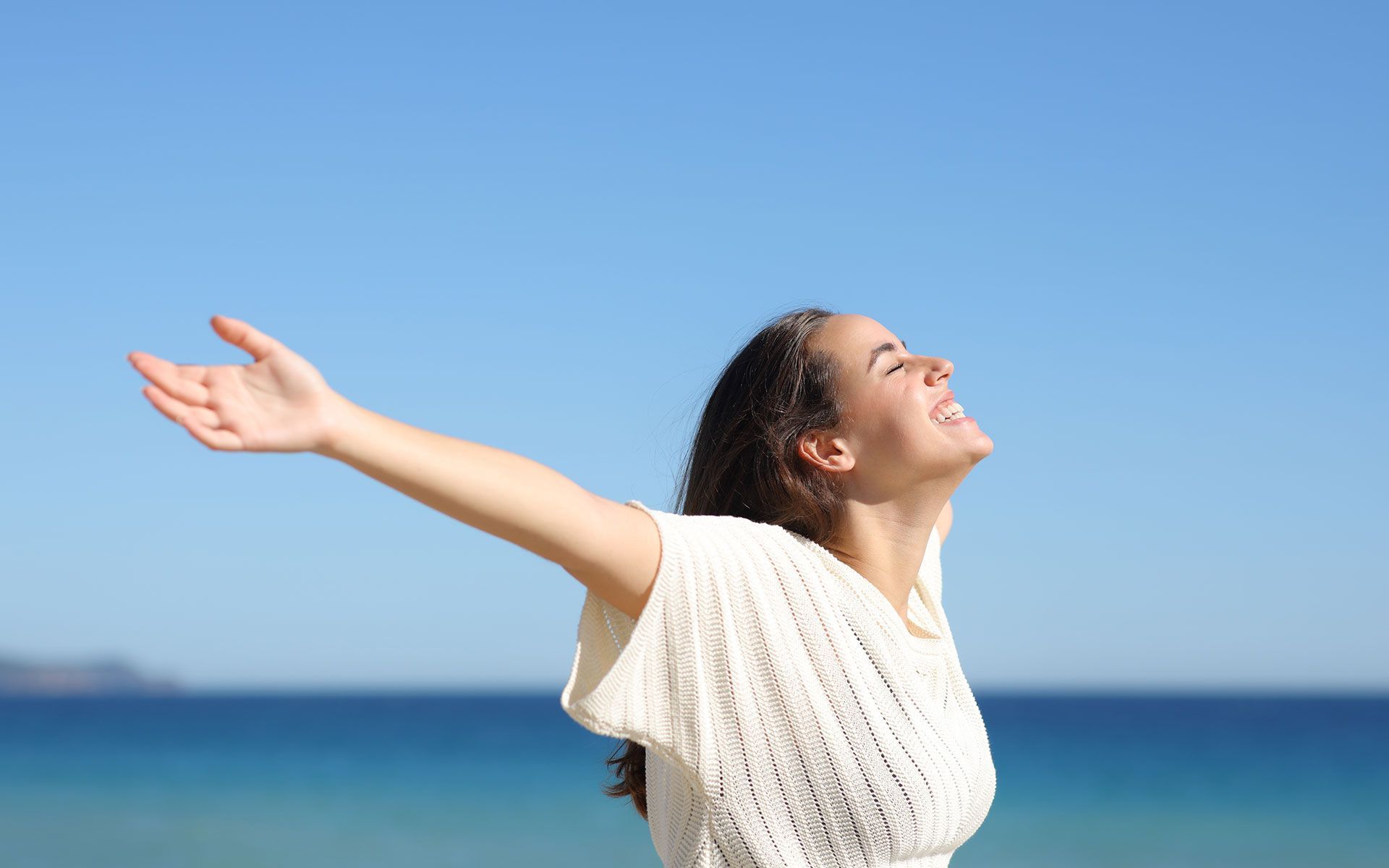 Woman with arms outstretched, smiling at the bright sky over a blue ocean.