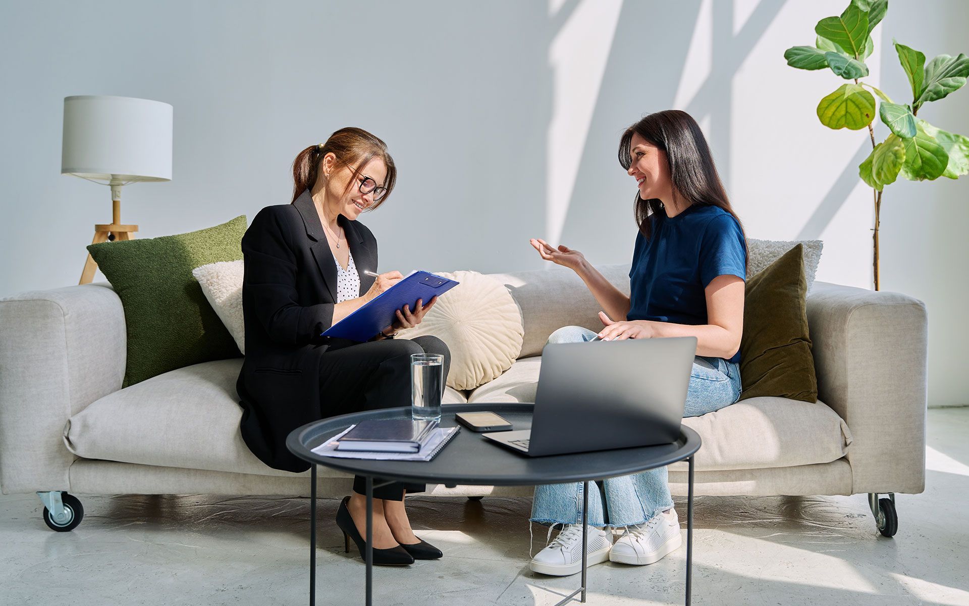 Woman in black suit takes notes as another woman gestures. They sit on a couch in a well-lit room.