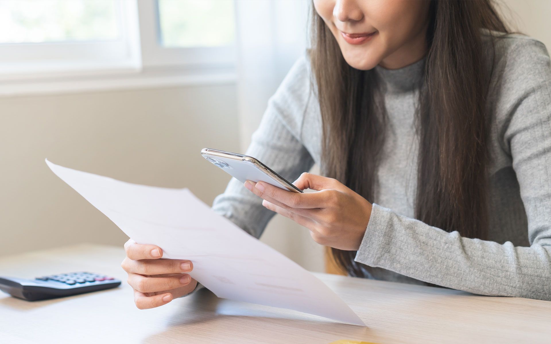 Woman scanning a document with a smartphone. Holding paper, smiling, and sitting at a desk indoors.