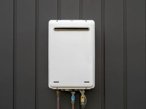 White outdoor tankless water heater mounted on a gray wall with pipes below