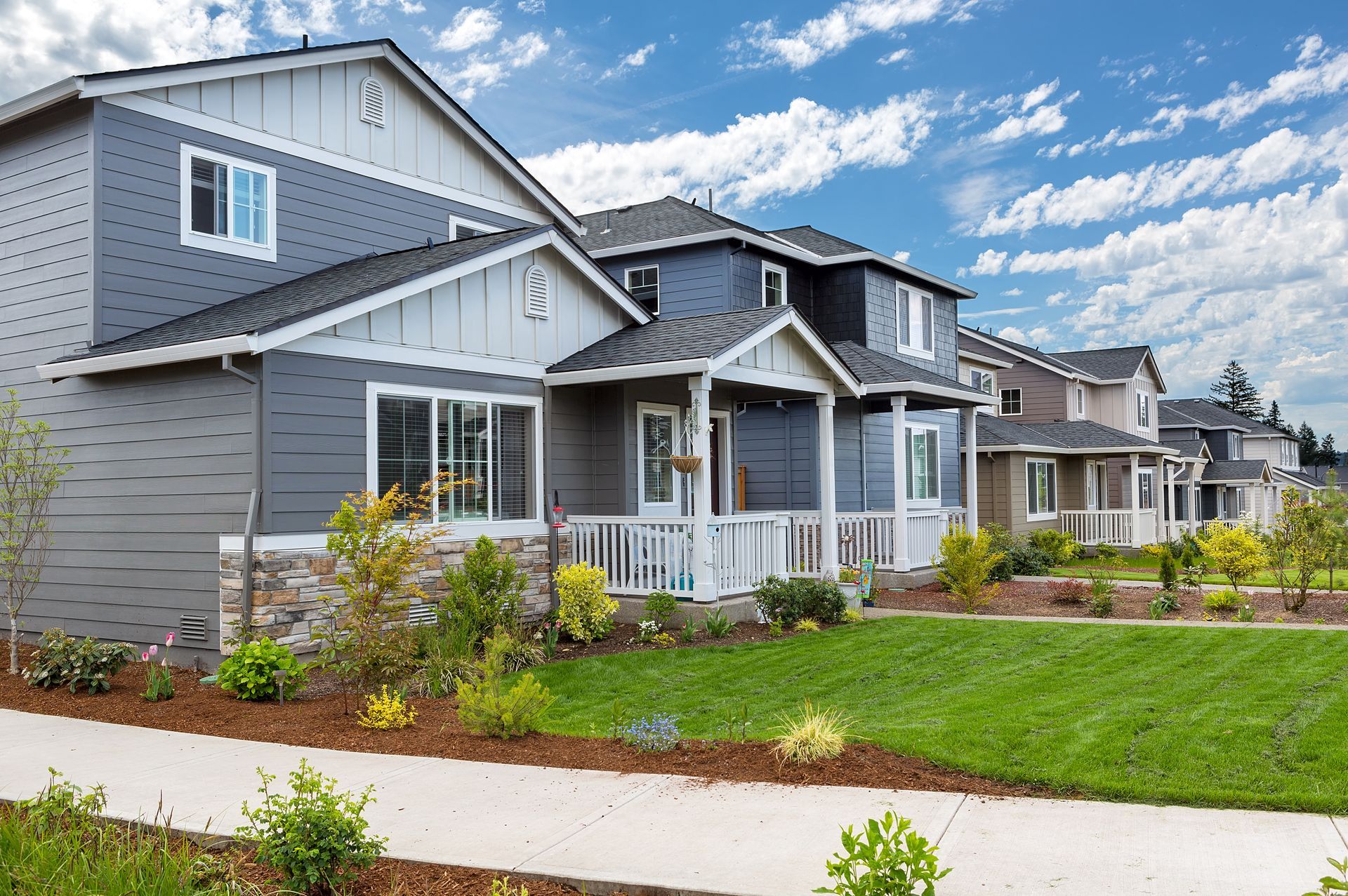 A row of modern, two-story homes with dark blue siding and stone accents under a bright, cloudy blue sky.