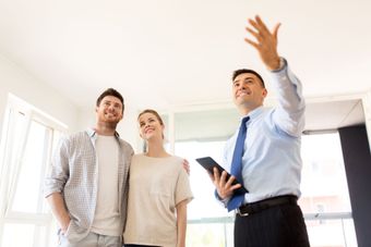 A real estate agent holds a tablet and gestures toward a ceiling for a smiling couple in a bright, unfurnished room.