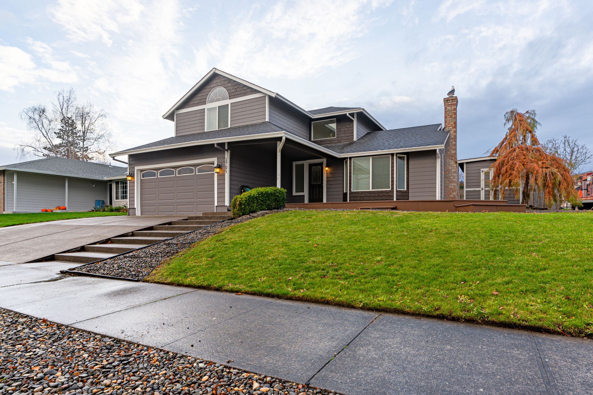 A two-story, gray-sided house with a stone chimney, a green lawn, and a wide driveway under a cloudy sky.