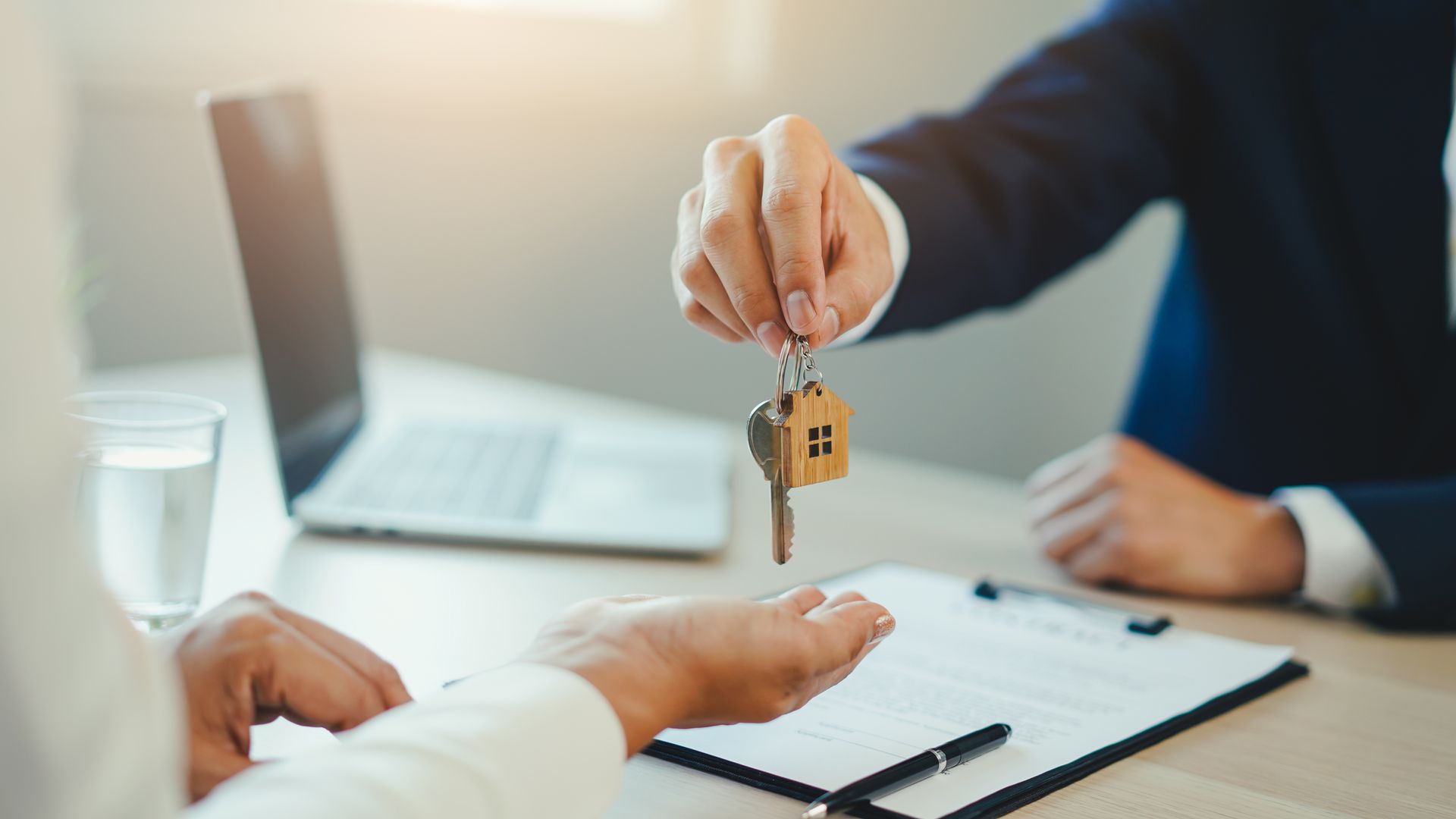 A person in a business suit hands keys with a house-shaped keychain to another person at an office desk.