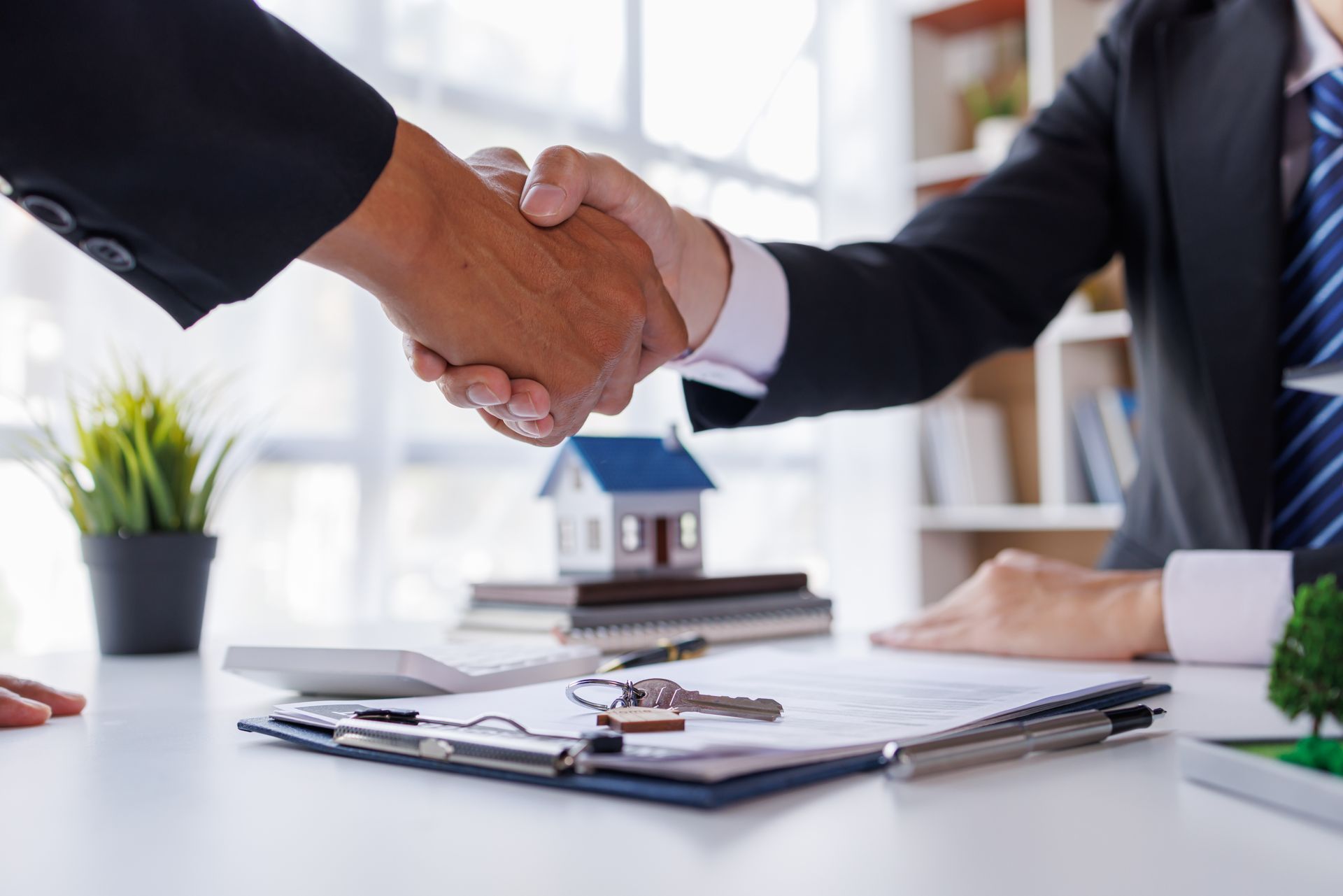 Two professionals in business suits shake hands over a desk with a small house model, documents, and keys.