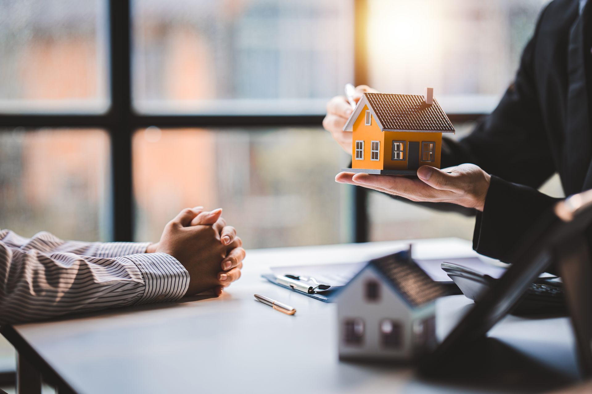A person in a business suit holding a small model house over a desk, with a client seated opposite.