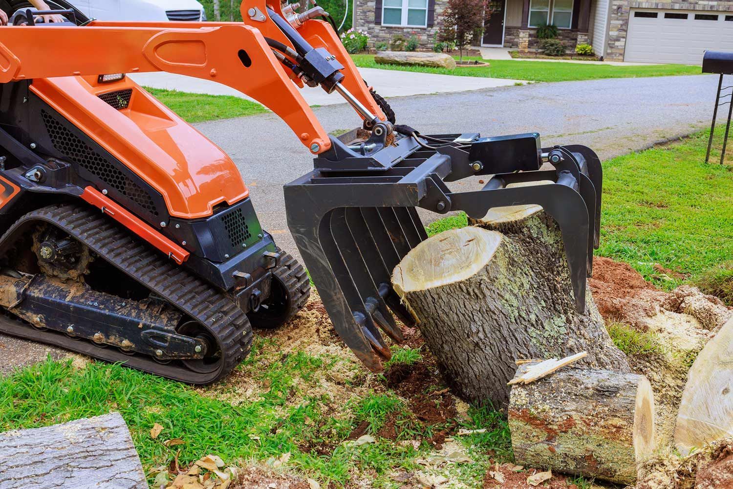 Mini excavator with grapple lifting a tree trunk on a lawn