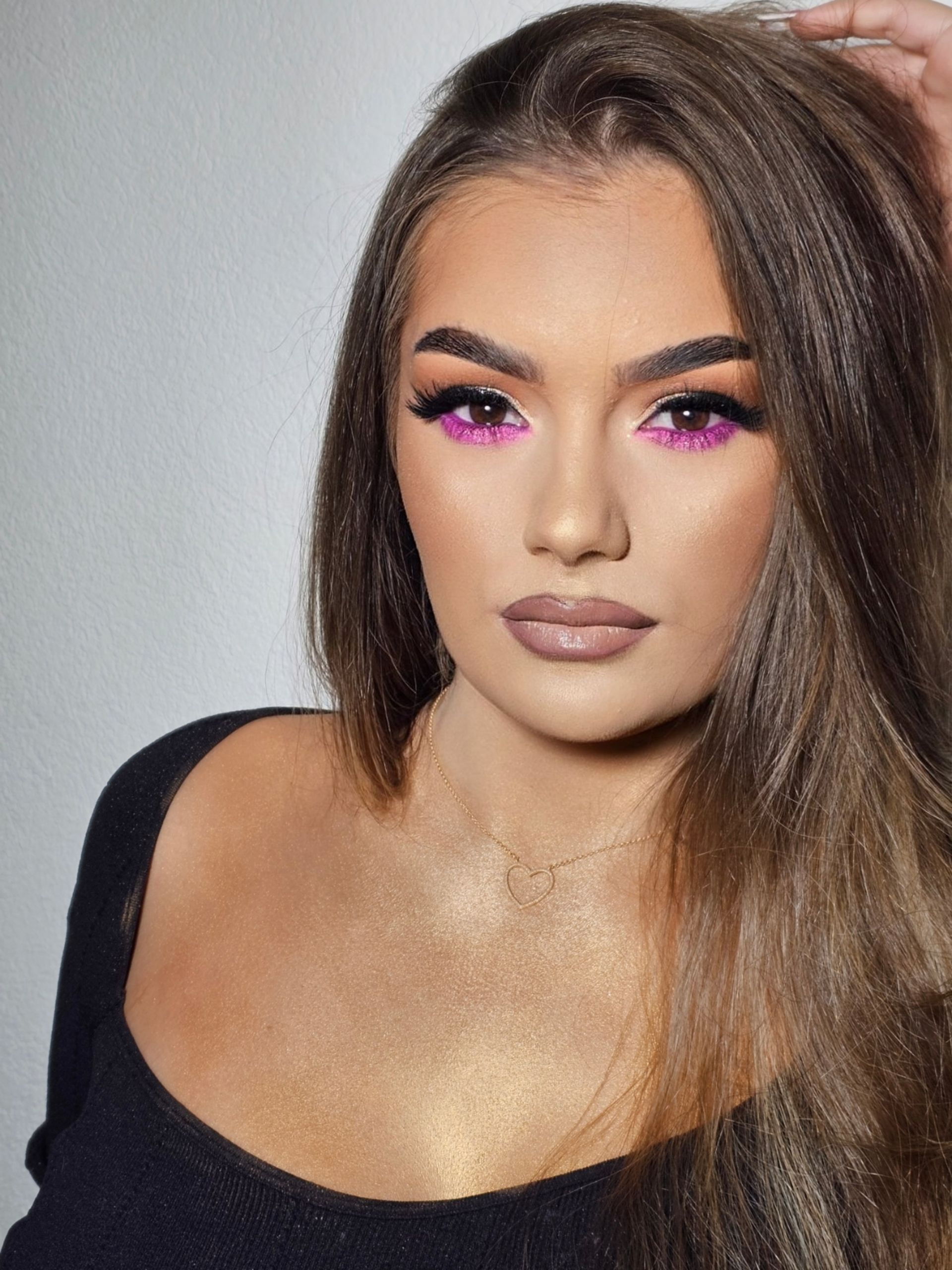 Frau mit braunem Haar, lila Augen-Make-up, schwarzem Oberteil und Goldglitter; posiert vor einer weißen Wand.