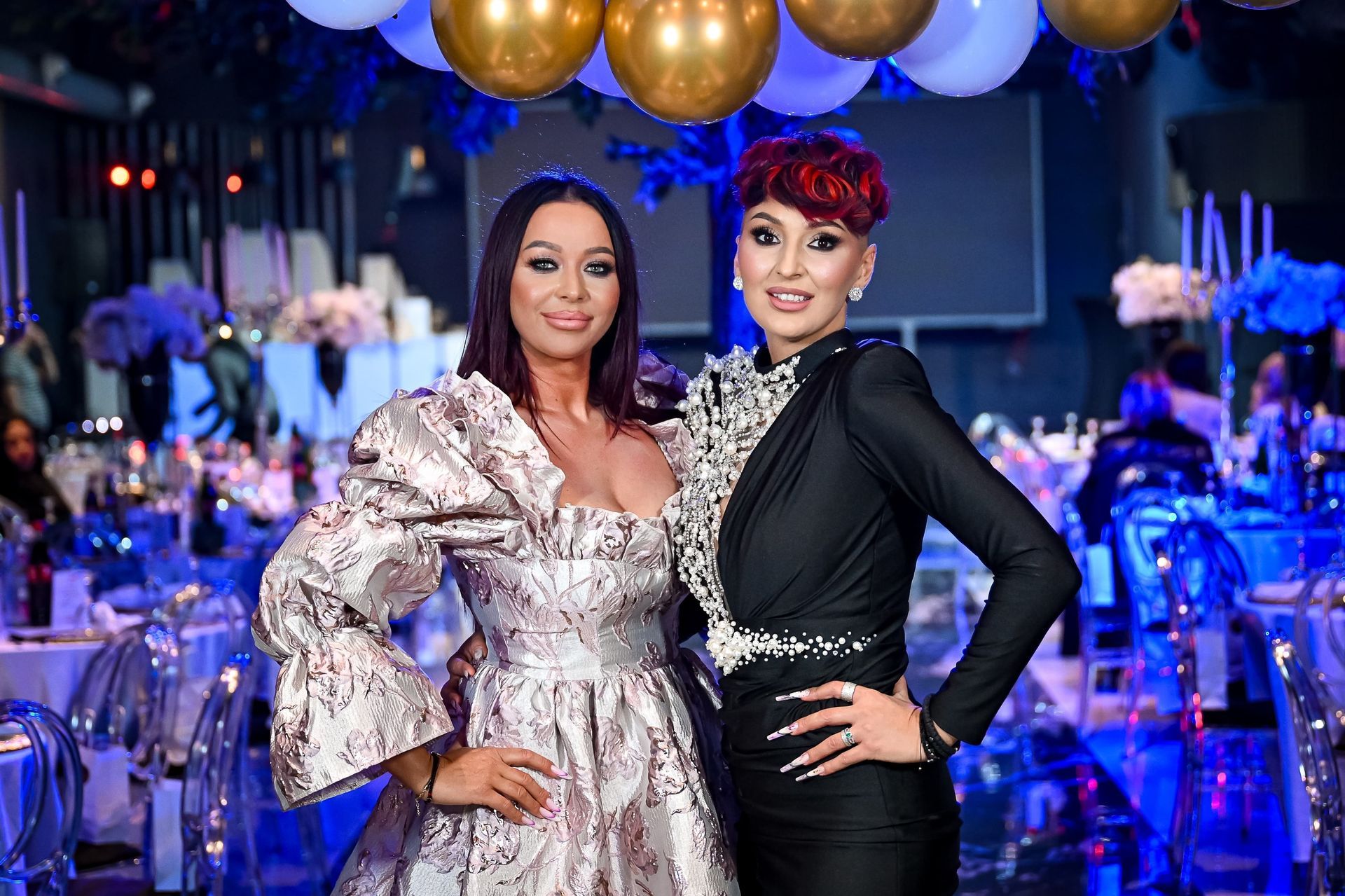 Zwei Frauen bei einer Veranstaltung, eine im gemusterten Kleid, die andere in Schwarz, beide lächelnd.