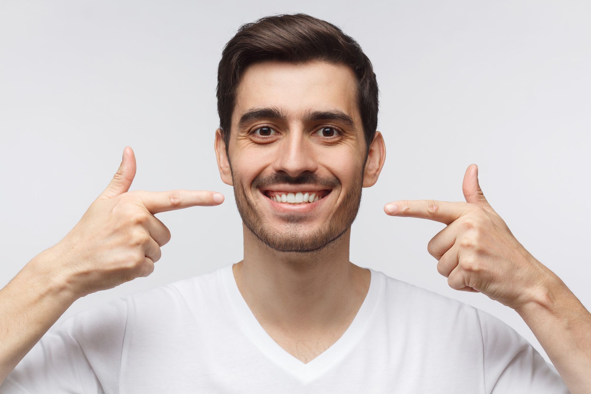 Man pointing at his smiling teeth, wearing a white shirt against a gray background.