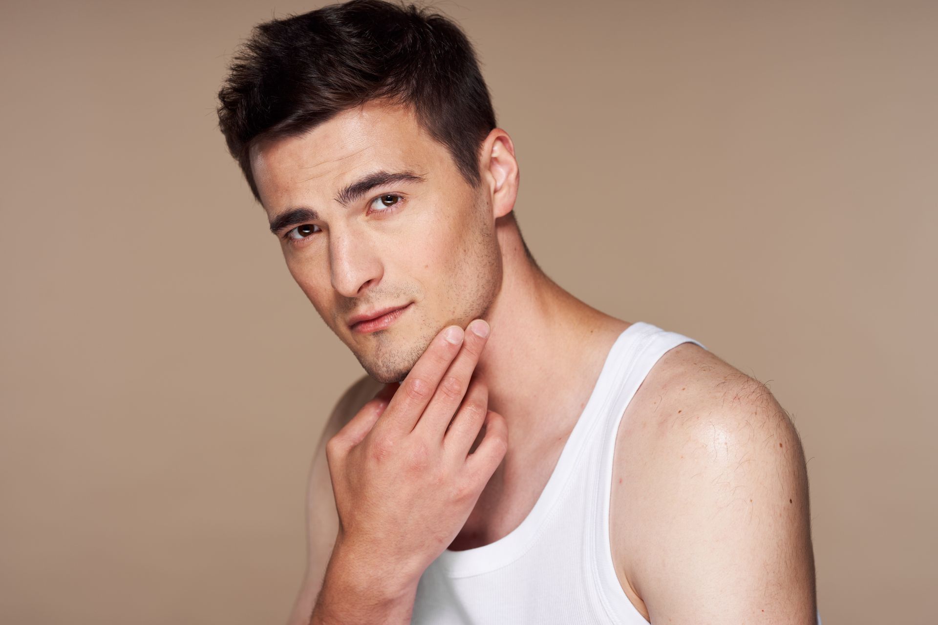 Man in white tank top touching his face, looking toward the camera. Beige background.