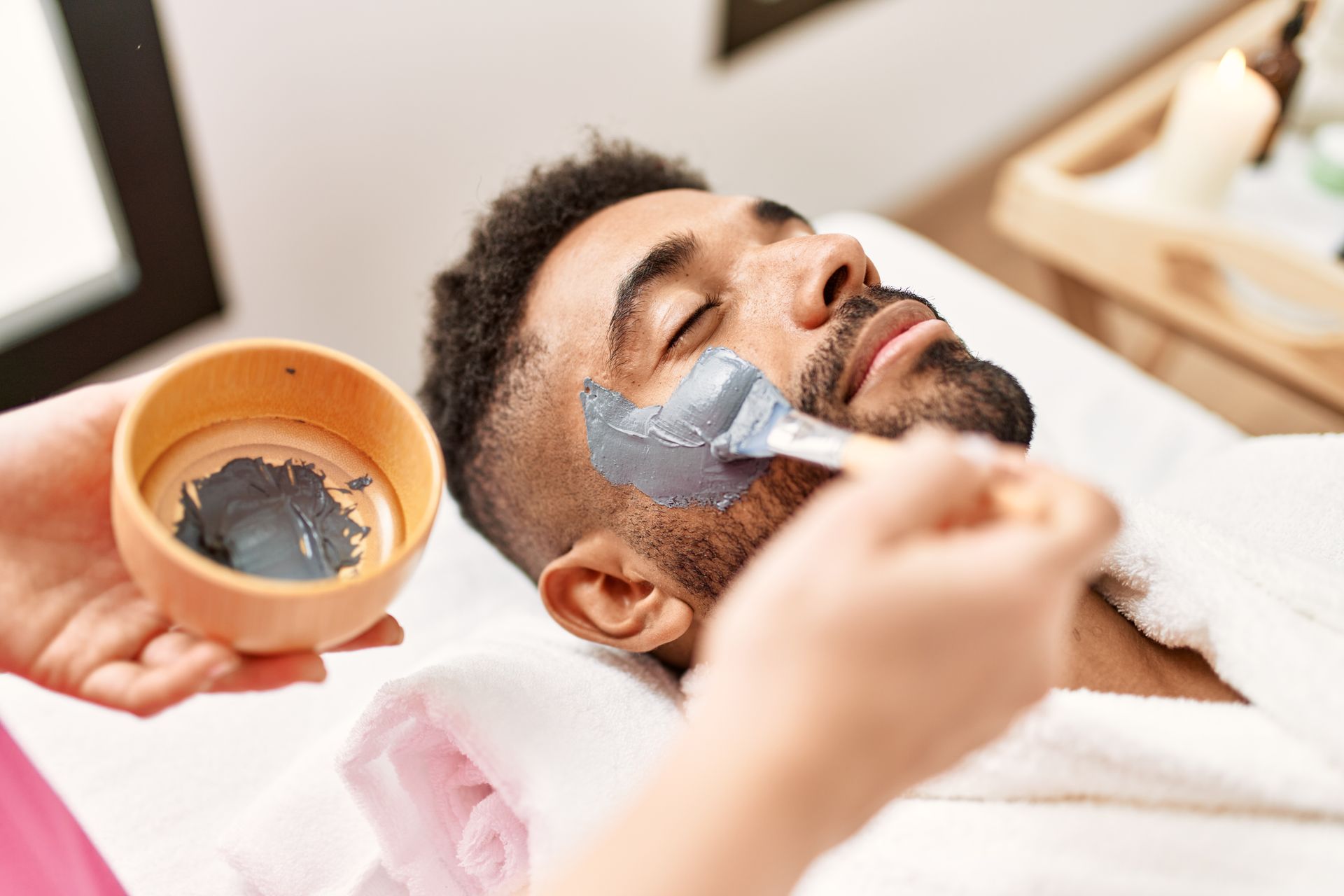 Person receiving a facial treatment with a gray mask applied with a brush.