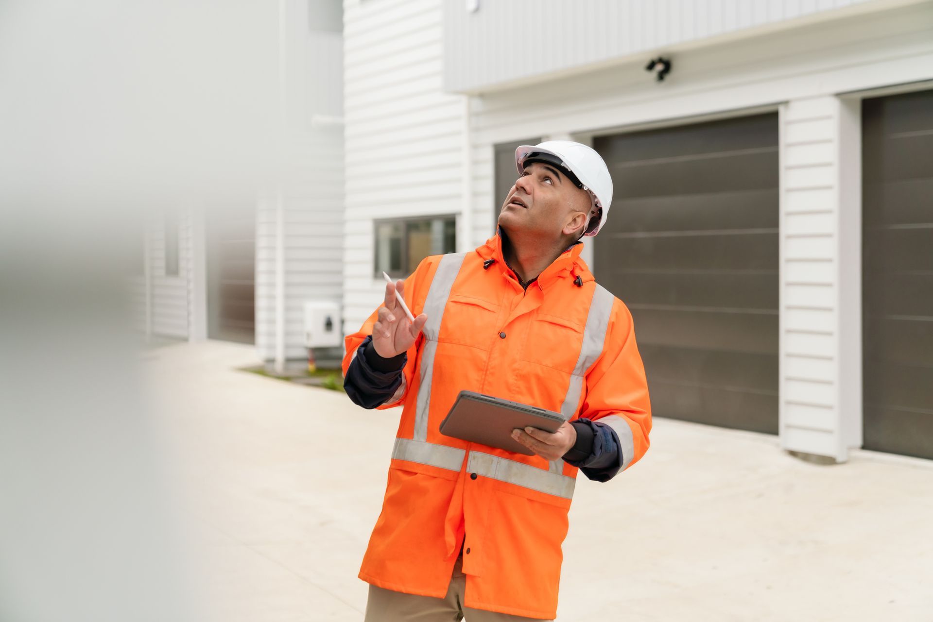 A man in an orange jacket and hard hat is standing in front of a house holding a clipboard.