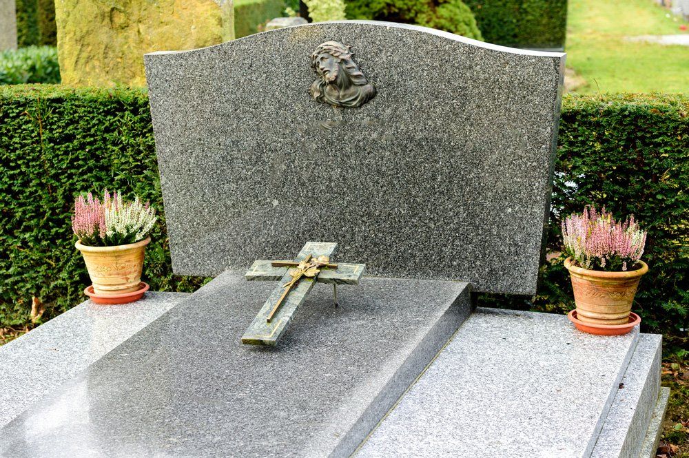 Lapide con crocifisso, affiancata da due piccoli vasi di fiori, in un giardino cimiteriale.