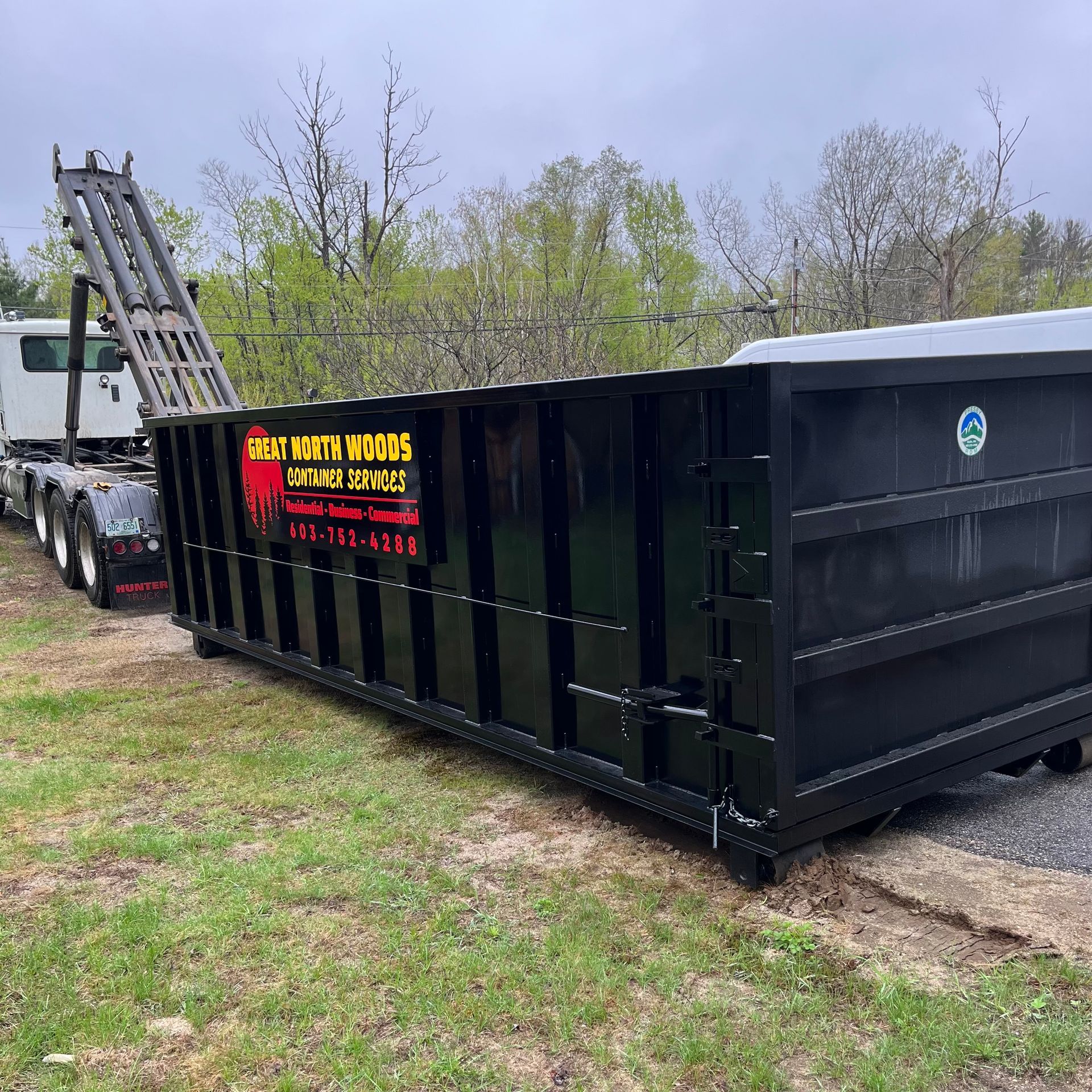 Black dumpster being loaded onto a truck on grassy terrain with trees in the background.
