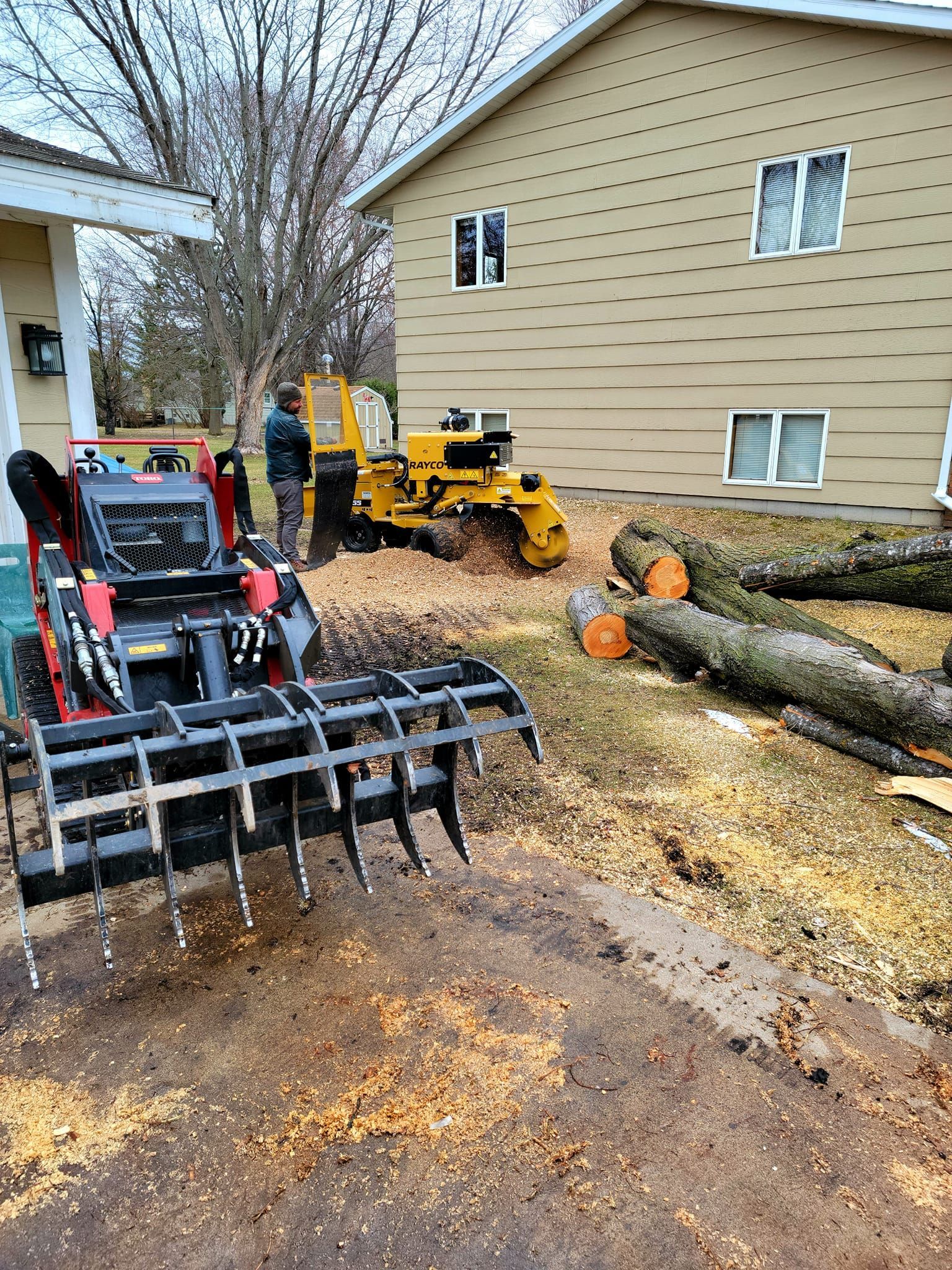 A compact track loader with grapple and a stump grinder working next to a house with cut logs.