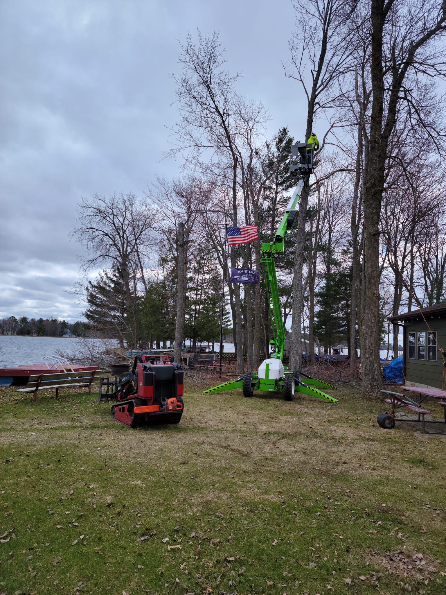 A person in a lift trimming a tree near a lake, red machine nearby. Green lift, gray sky, autumn setting.