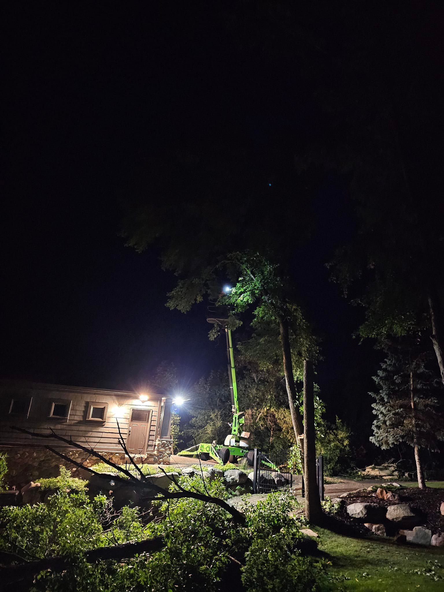 Night scene of tree trimming; a large green lift with workers, bright lights illuminating branches and building.