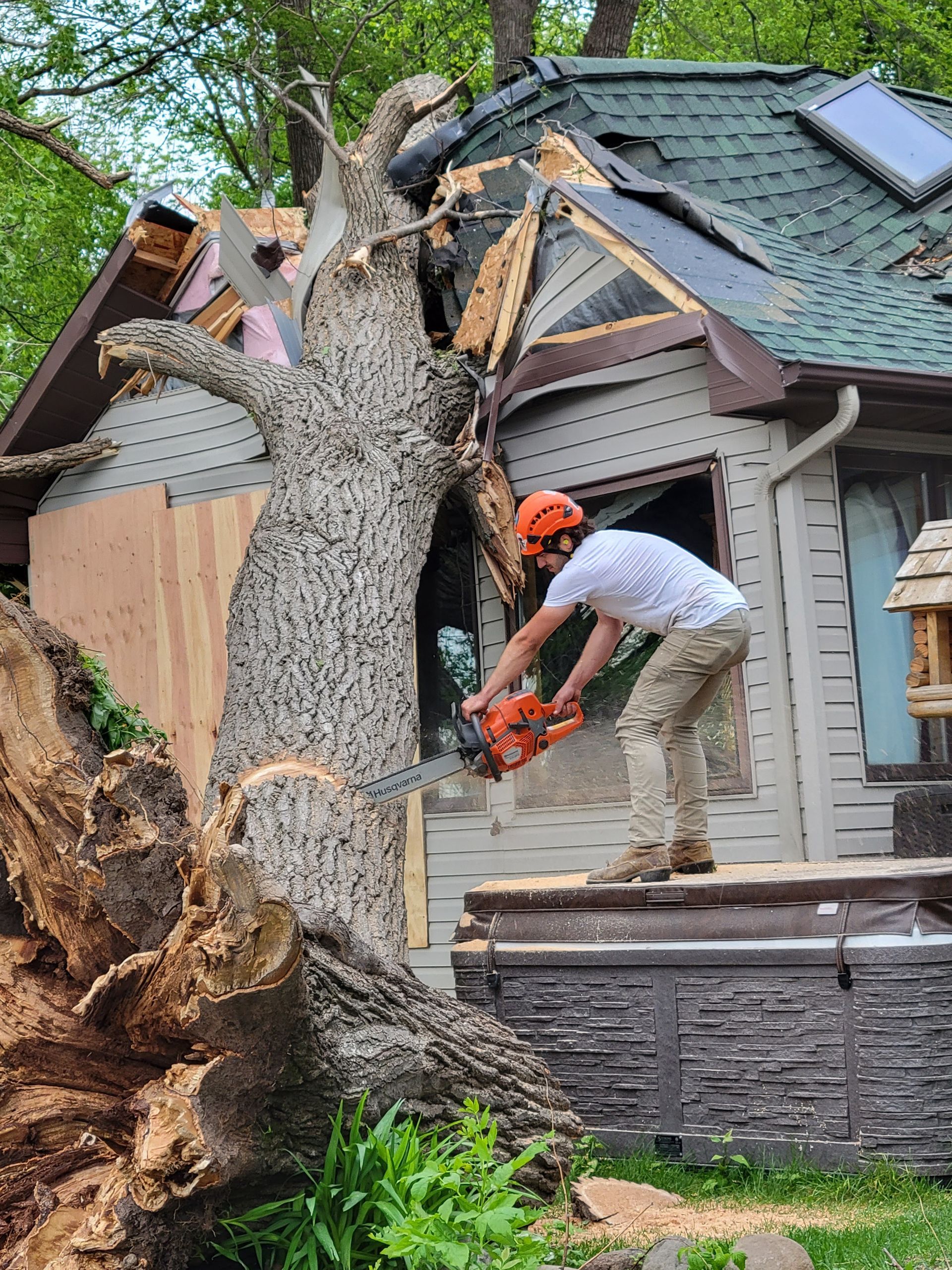 Man in hardhat cutting a tree lodged in a house with a chainsaw. The house roof is damaged.